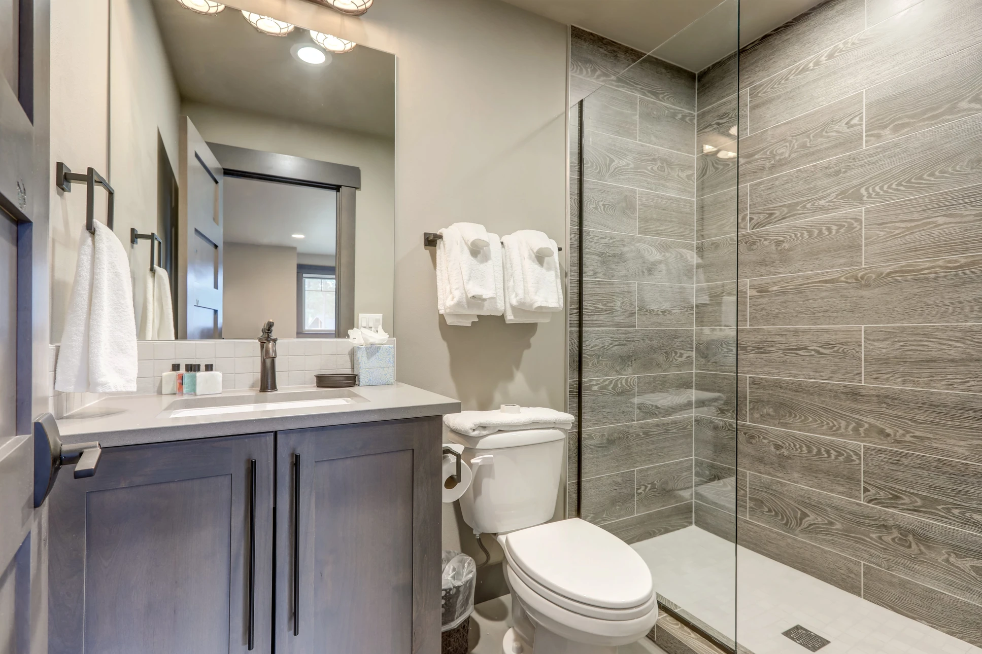 Salle de bain moderne avec douche à l'italienne aux carreaux effet bois gris, meuble-vasque en bois foncé avec plan en quartz blanc, miroir encadré noir, toilettes blanches et accessoires coordonnés