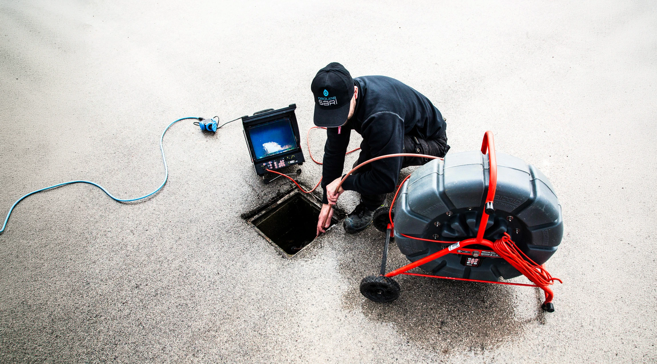 Technicien en combinaison noire accroupi sur une surface en béton, inspectant un regard d'égout ouvert