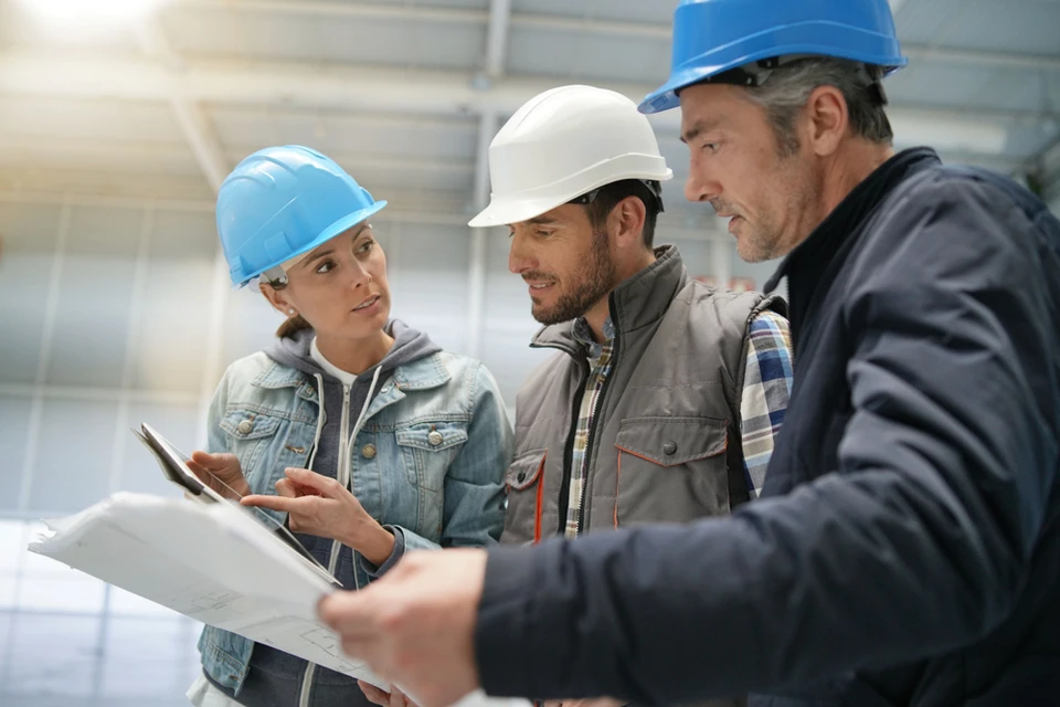 Réunion de chantier en intérieur avec trois professionnels du bâtiment portant des casques de sécurité colorés, consultant des plans architecturaux