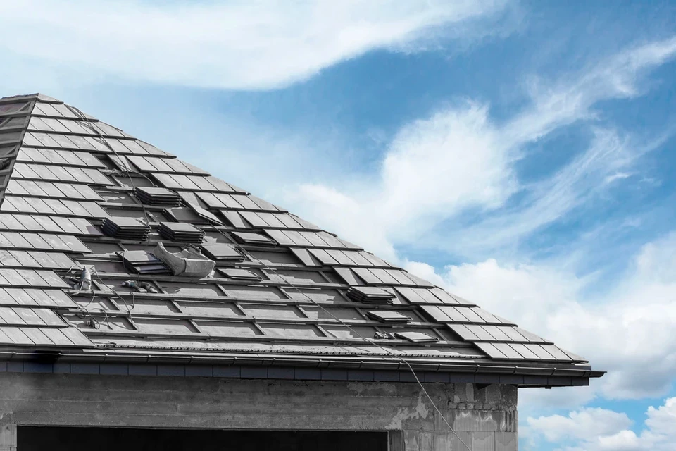 Ouvrier couvreur en tenue de protection travaillant sur toiture en tuiles mécaniques grises, ciel bleu dégagé
